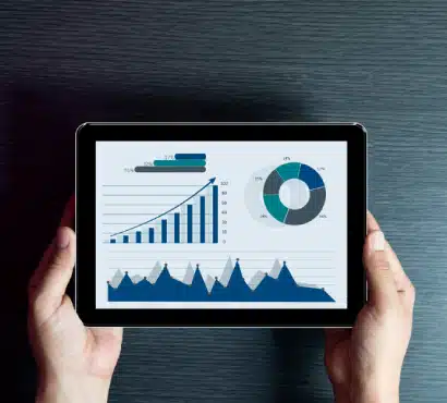 A person holding a tablet displaying various data visualizations, including a bar chart, a pie chart, and a line graph with blue and green colors, against a dark wood background.