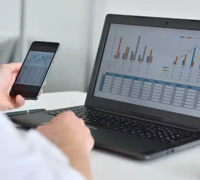 A person is working at a desk, holding a smartphone displaying charts and looking at a laptop screen with colorful graphs and data tables.