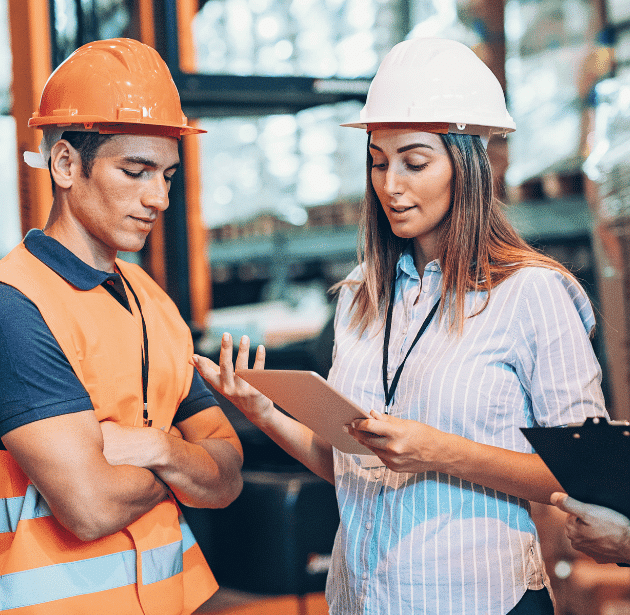 Two workers wearing safety helmets and vests are standing in a warehouse. The woman is holding a digital tablet and pointing at the screen, while the man listens attentively with his arms crossed.