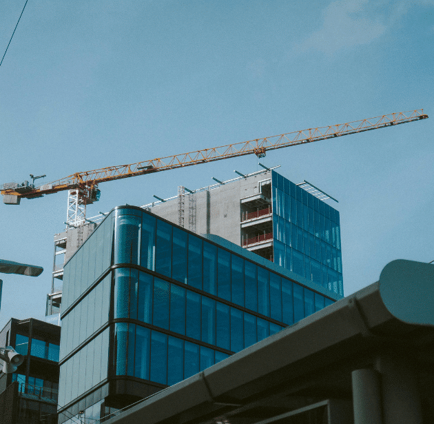 A yellow construction crane stands over a modern glass office building under construction, with blue skies in the background and parts of other nearby structures visible in the foreground.