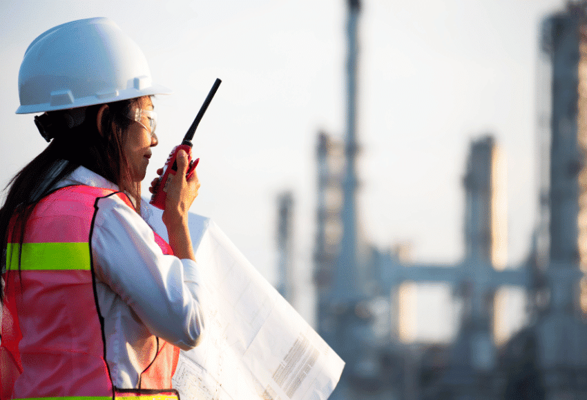 A person wearing a hard hat and reflective safety vest holds blueprints and speaks into a walkie-talkie at an industrial site with blurred structures in the background.