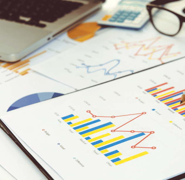A close-up of financial charts and graphs on paper, showing bar and line graphs with various colored data points, next to a laptop keyboard and a pair of glasses.