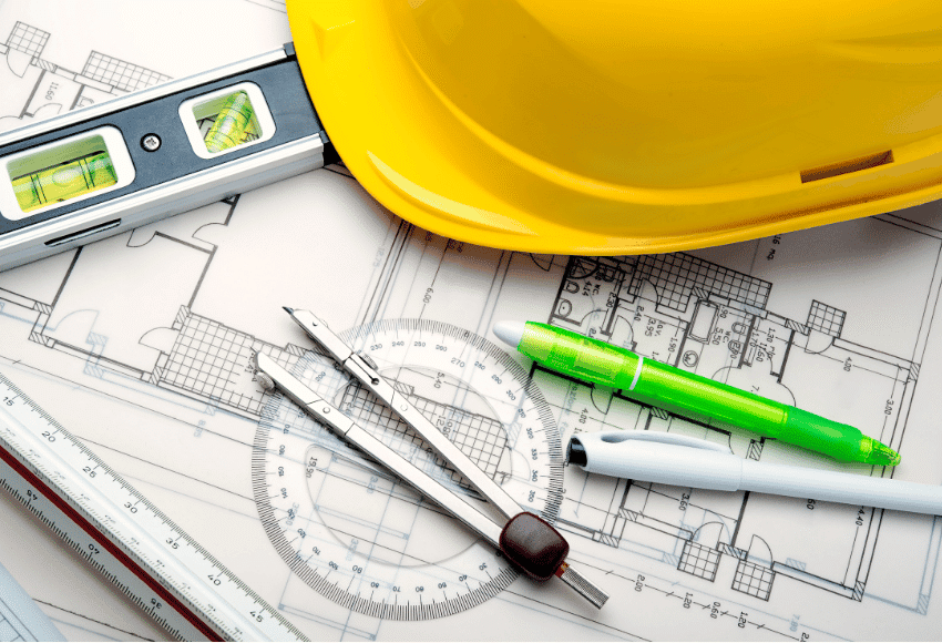 A yellow hard hat, level tool, compass, protractor, ruler, green pen, and black marker are placed on top of architectural blueprints.