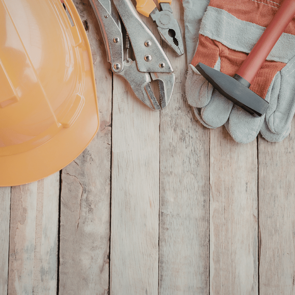 Yellow hard hat, pliers, screwdriver, work gloves, and a wrench laid out on a light wooden surface, representing construction or maintenance tools and safety equipment.