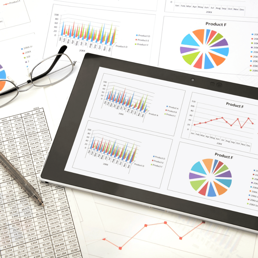 A tablet displays colorful bar and pie charts, surrounded by printed charts, graphs, financial spreadsheets, a pen, and a pair of glasses on a white desk.